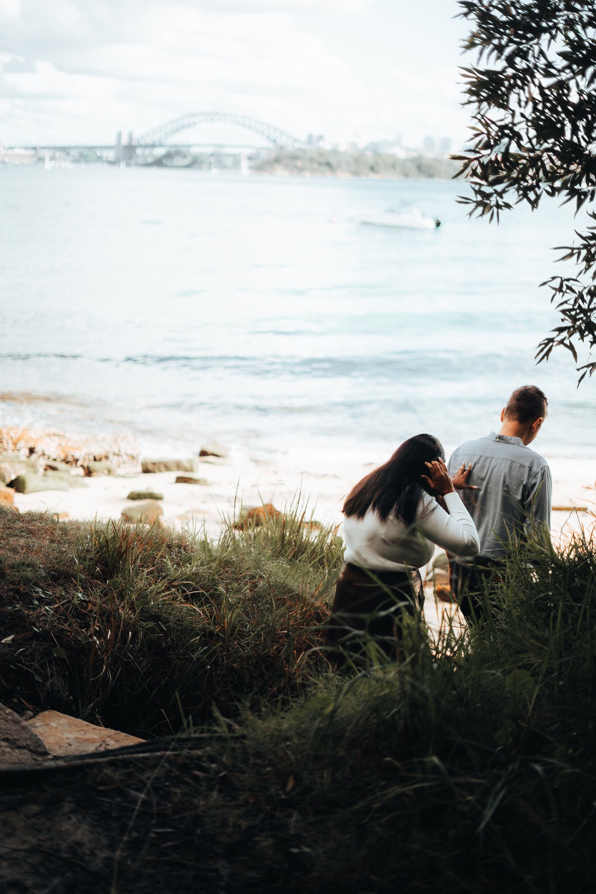 A couple sitting on a grassy shoreline overlooking a body of water, with a bridge and distant city skyline in the background. The photo is framed by tree branches at the top right, and the couple is seen from behind.