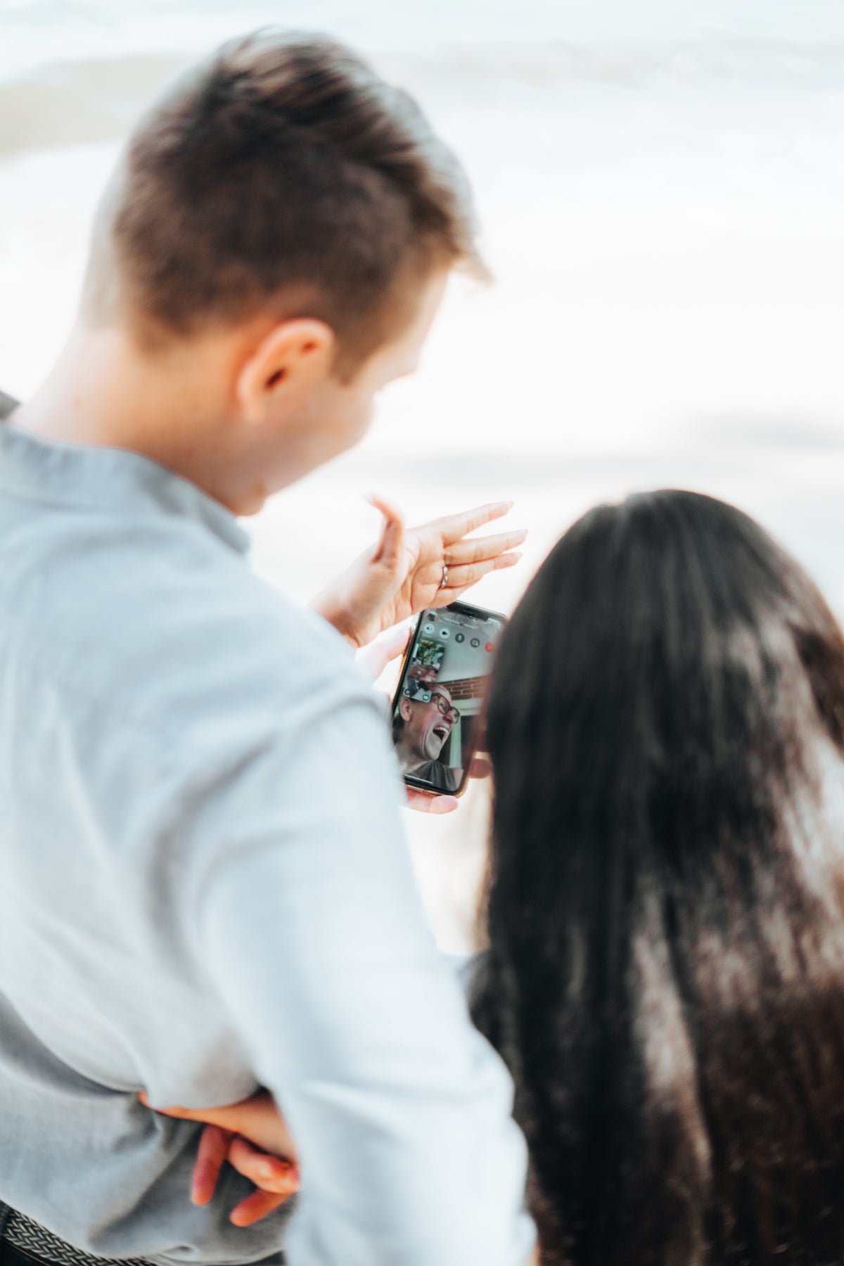 A man in a light blue shirt and a woman with long dark hair are seen from behind, looking at a smartphone screen together in an outdoor setting with a bright, soft-focus background.