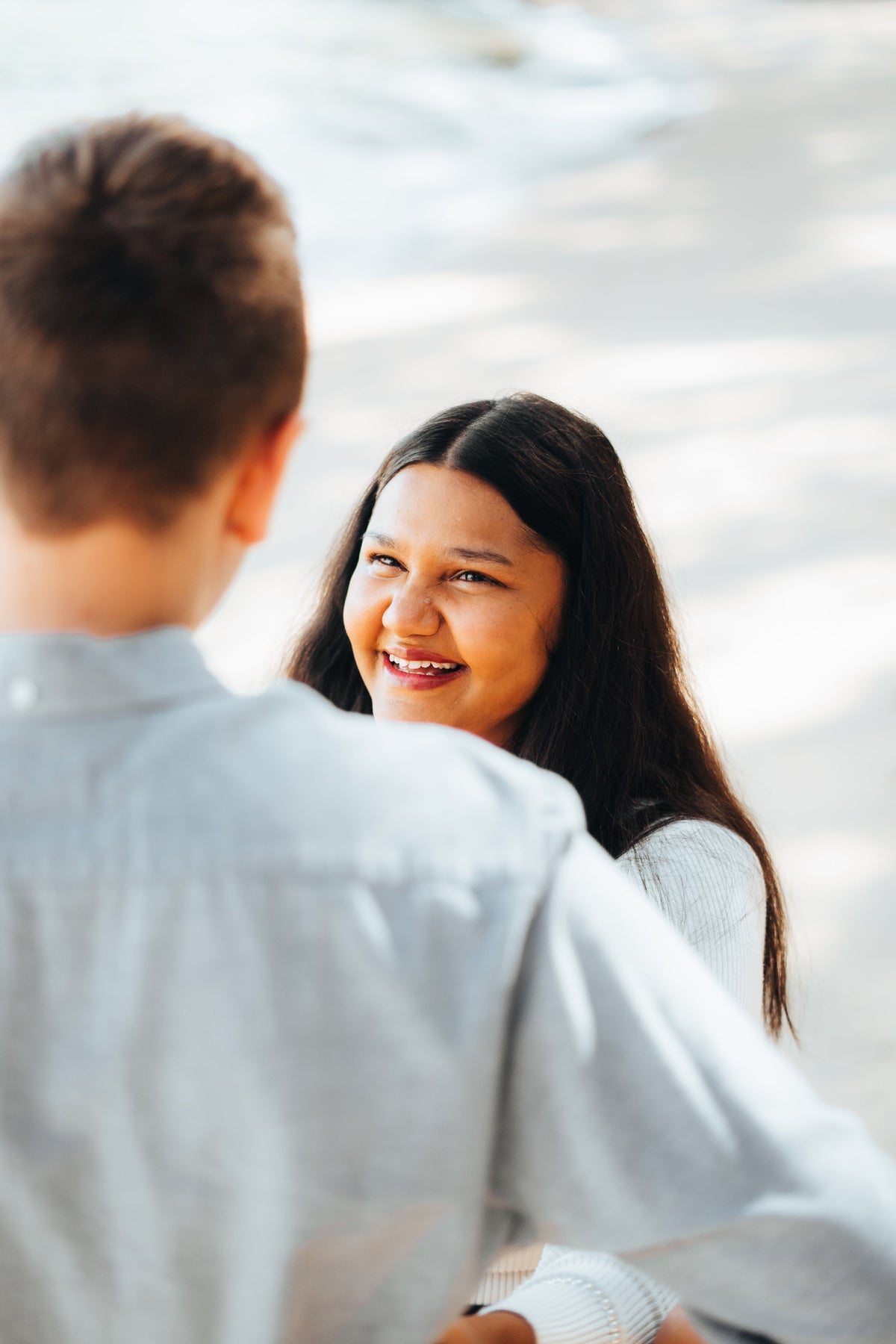 A smiling woman with long dark hair looks up and engages in conversation with a man whose back is turned, photographed outdoors in soft natural light.
