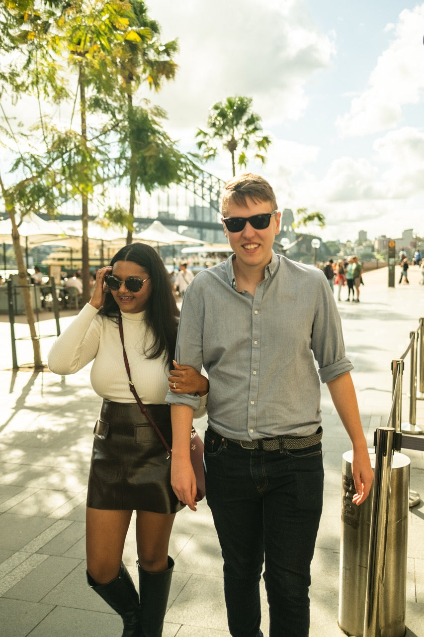A man and a woman walking arm-in-arm outdoors on a sunny day. The man wears a light gray button-down shirt, black jeans, and sunglasses, while the woman wears a white long-sleeve top, a dark brown leather skirt, and black knee-high boots.