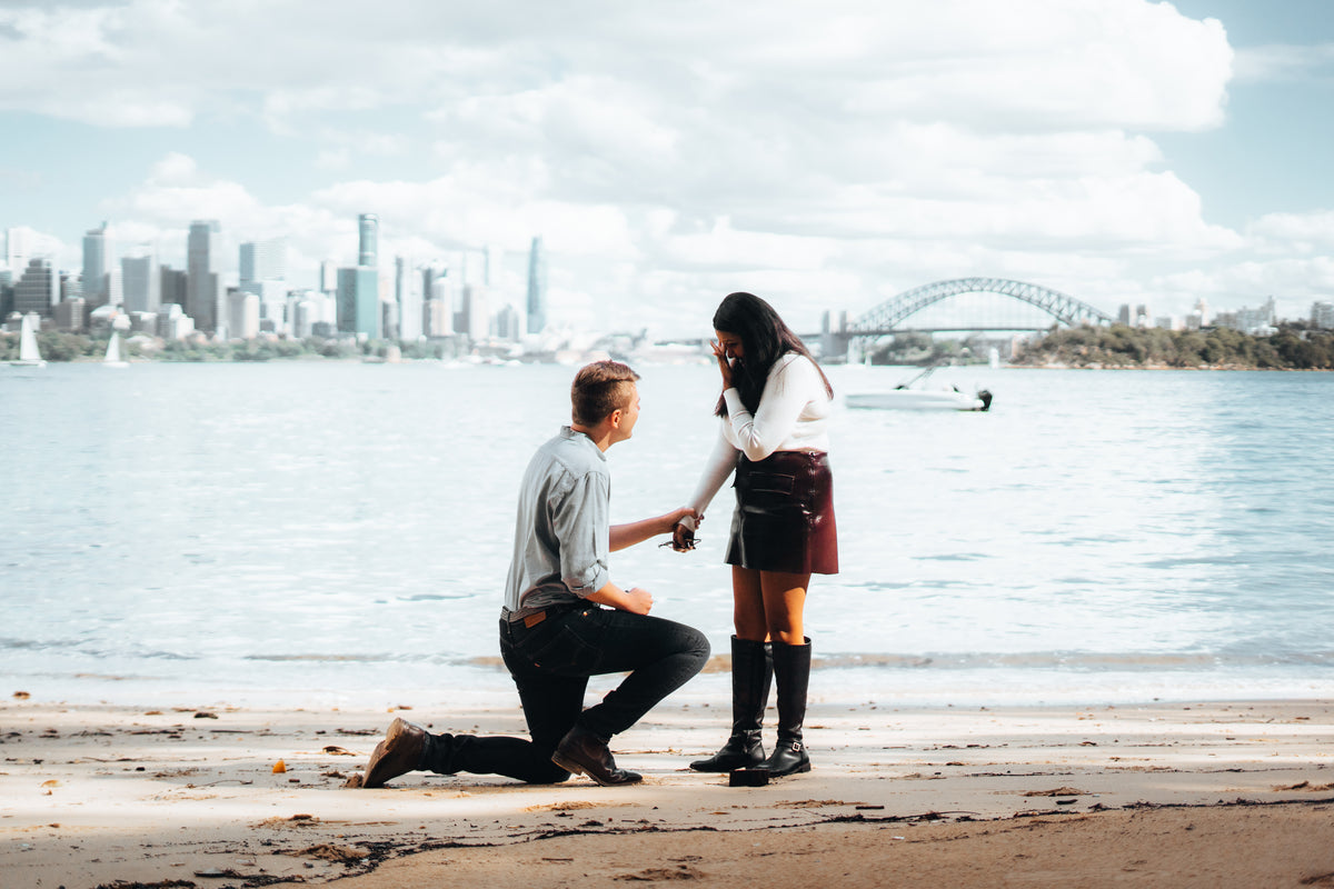 A man kneels on a sandy beach holding a ring box to a woman standing on the shore, with a wide ocean, city skyline, and bridge in the background.
