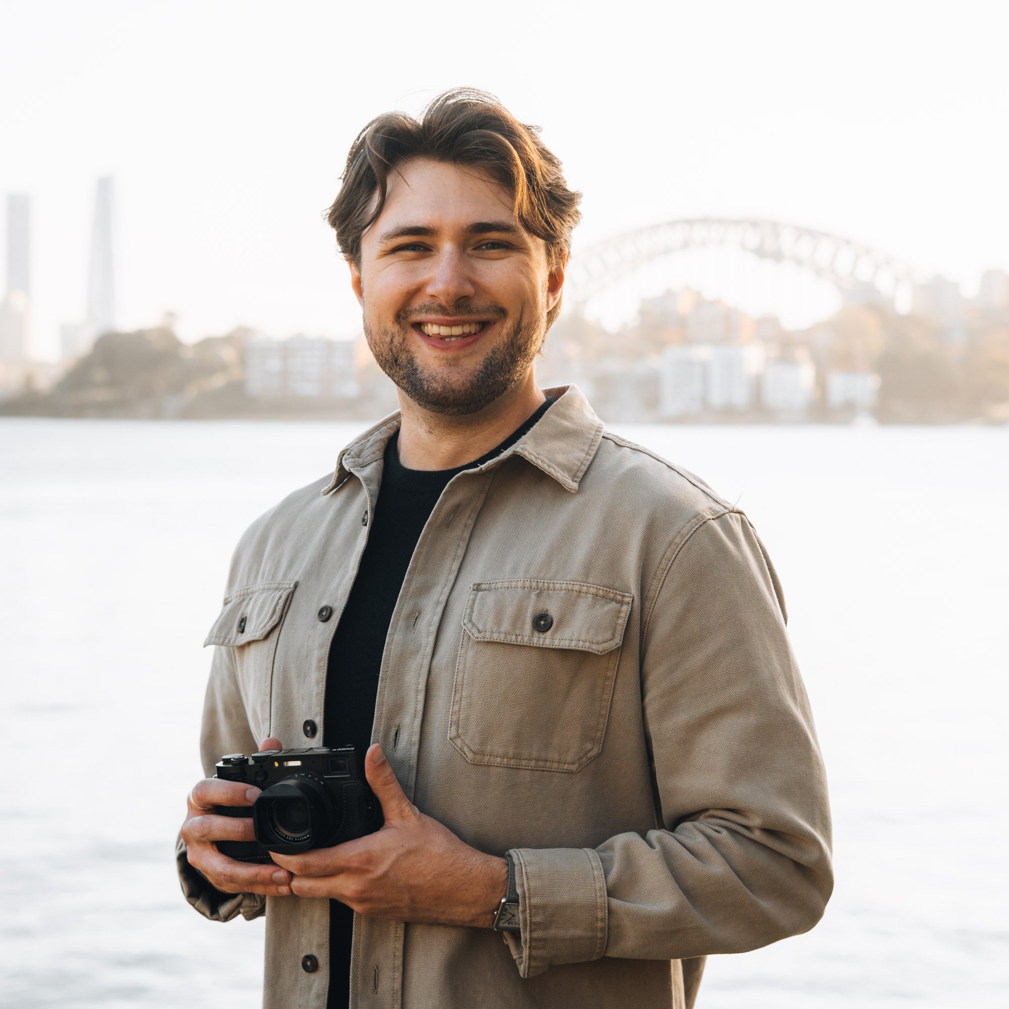 A smiling man wearing a beige overshirt over a black t-shirt, holding a black camera, with a soft-focus waterfront cityscape in the background.